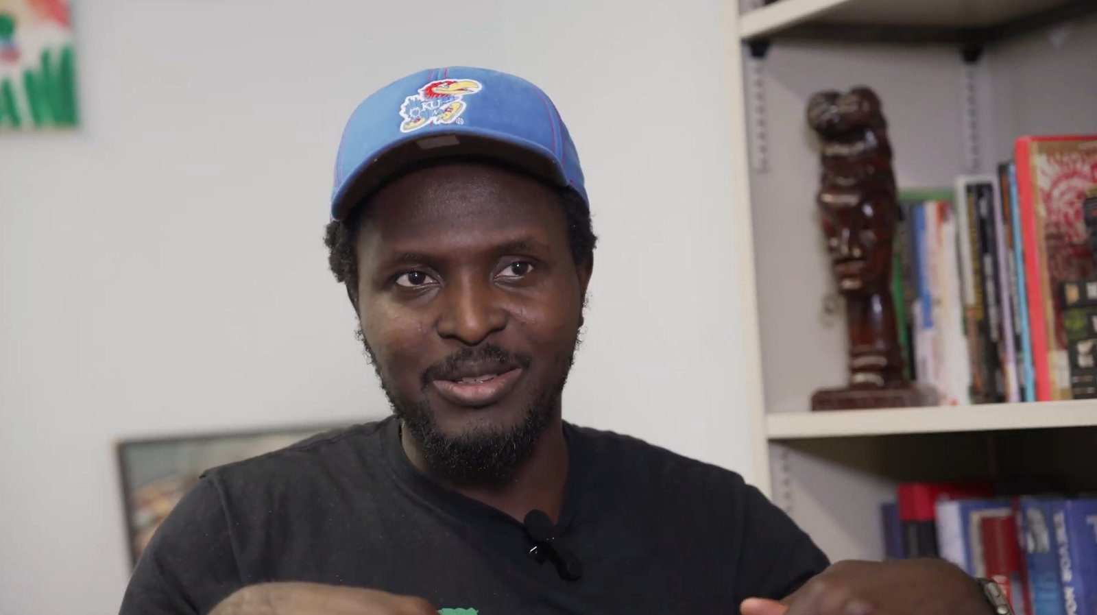 James Yeku seated in front of bookshelves during his filmed interview, wearing a Kansas cap